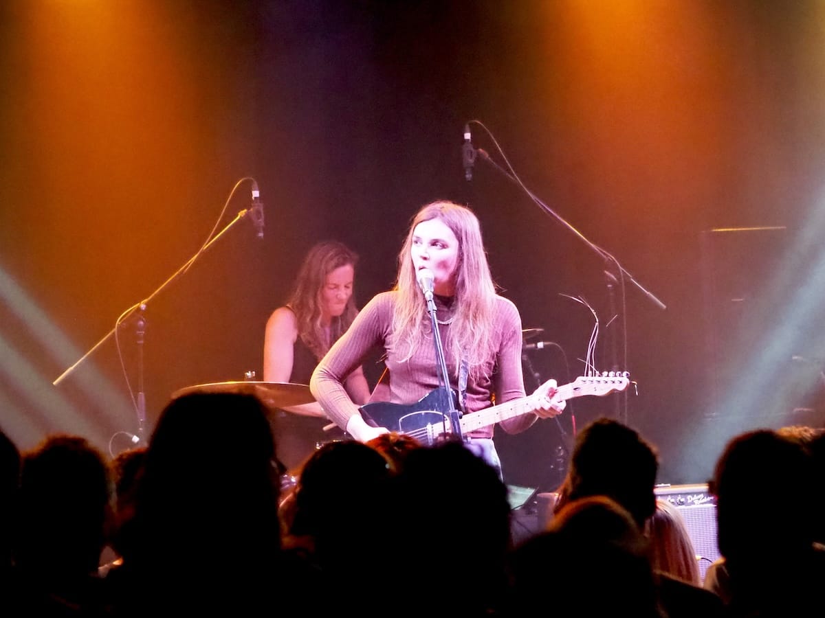 Romy Vager on stage playing guitar with the drummer visible in the background.