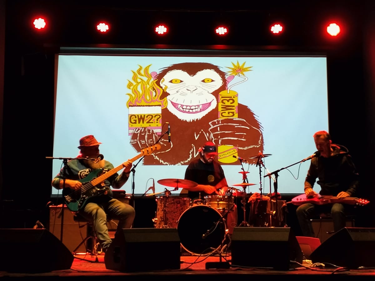 A three-piece band playing in front of a projected image of a monkey.